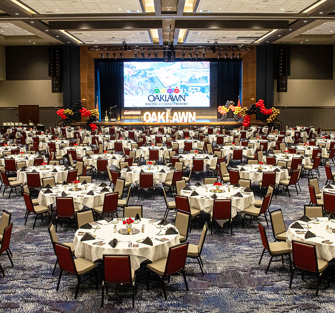 A vast room filled with finely set dinner tables leads to a stage with instruments and a theater-style screen